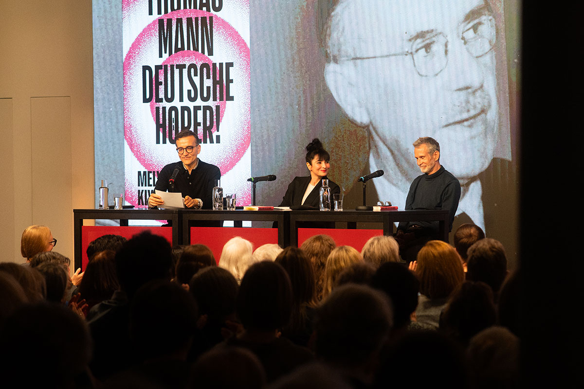 Daniel Schreiber mit Mely Kiyak und Schauspieler Ulrich Matthes auf der Bühne des Literaturhauses. Im Hintergrund ein Bild von Thomas Mann